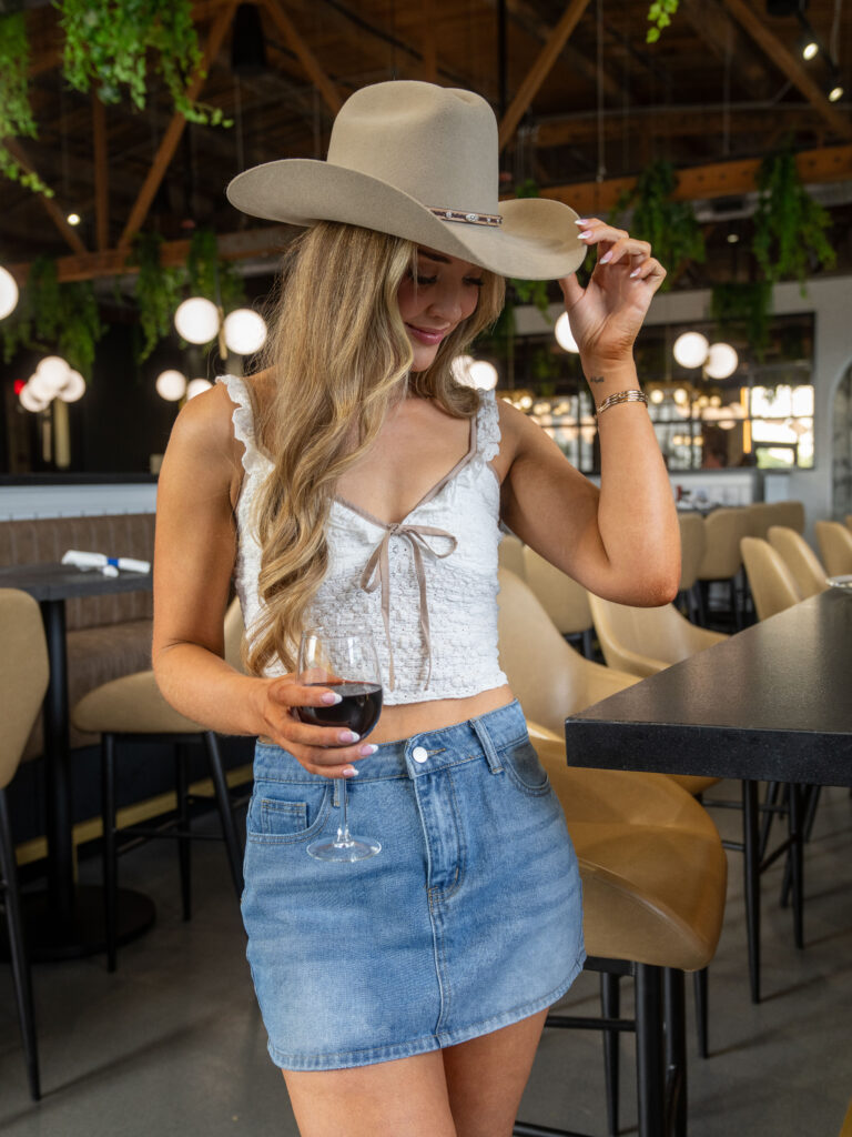 Woman wearing a western hat and holding a glass of wine inside a modern restaurant.